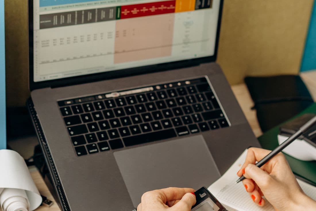 A laptop showing a colour coded spreadsheet while a hand writes notes in a notebook beside it