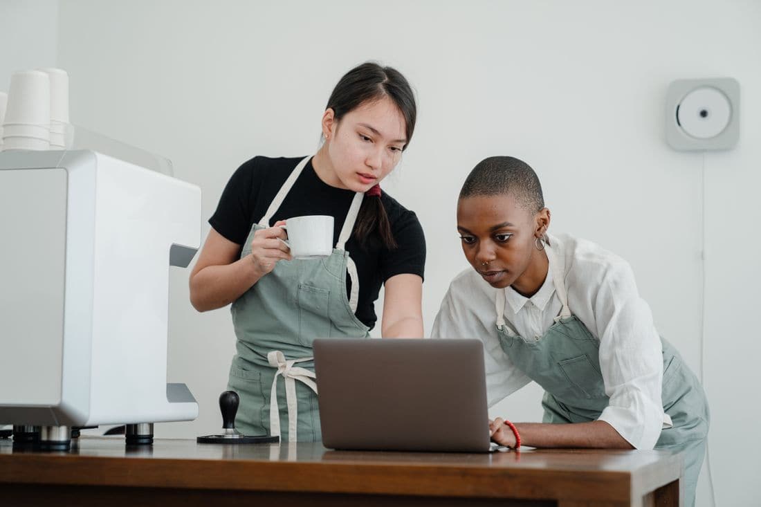 Two cafe workers in aprons checking a laptop on the counter beside the coffee machine