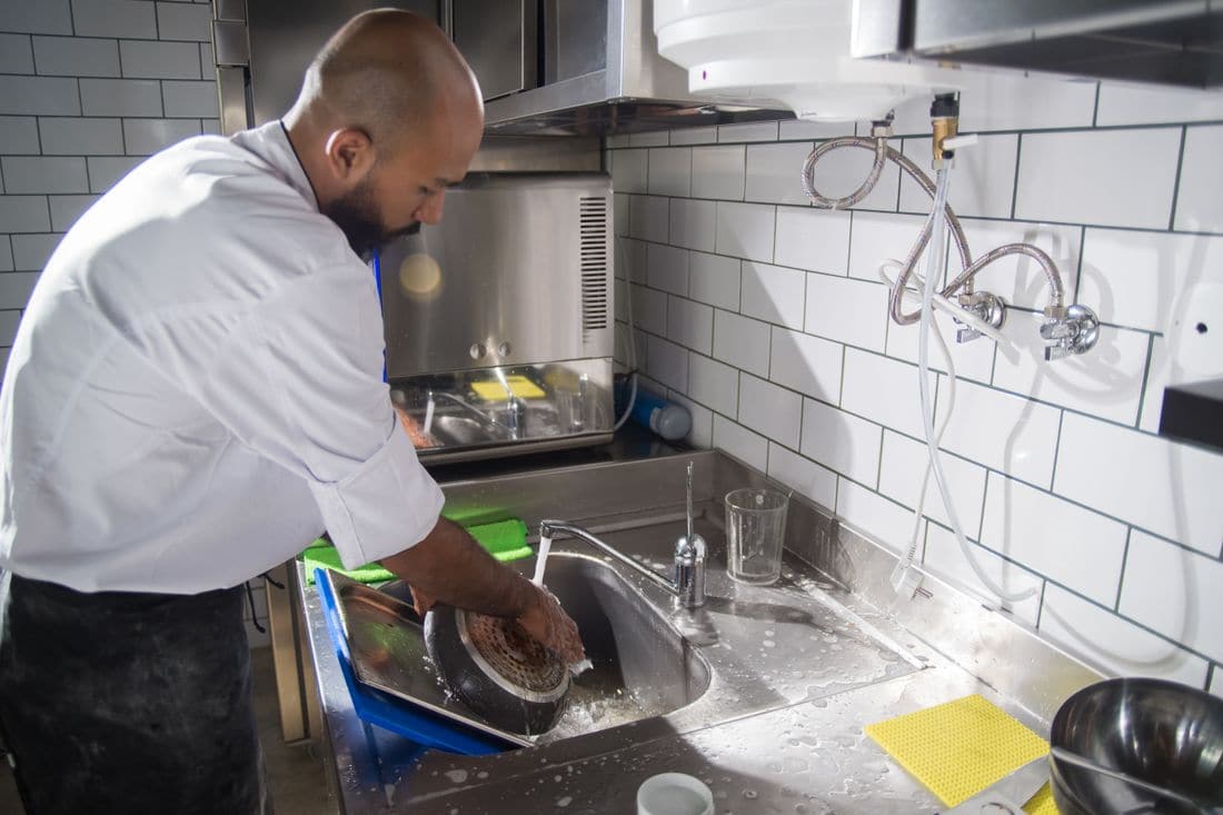 A cook washing a pan at a stainless steel sink in a commercial kitchen