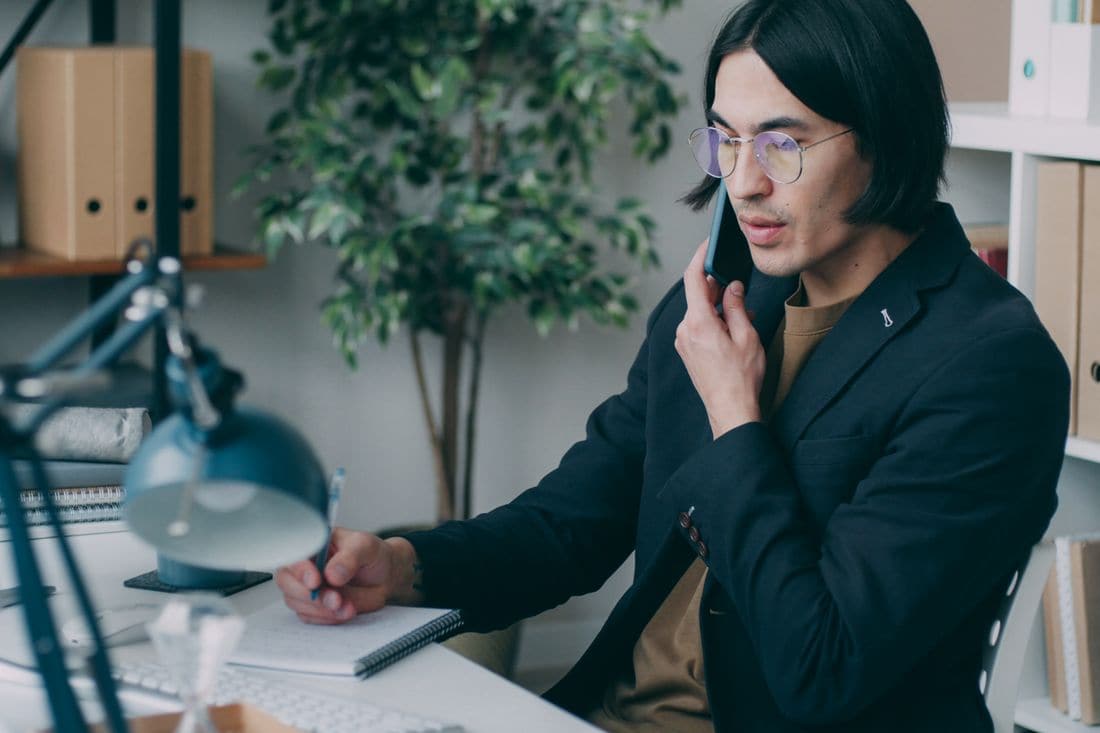 A man in glasses on the phone making notes at a desk
