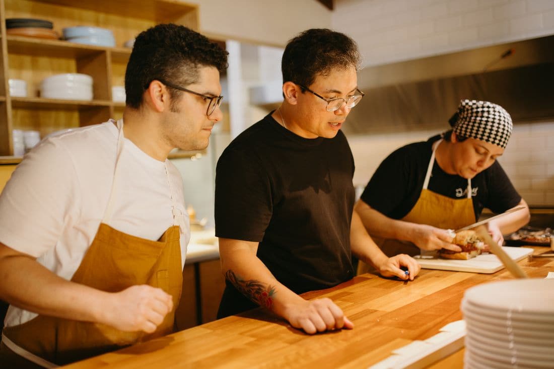 A cook in a black T-shirt talking two colleagues in aprons through a task at the kitchen counter