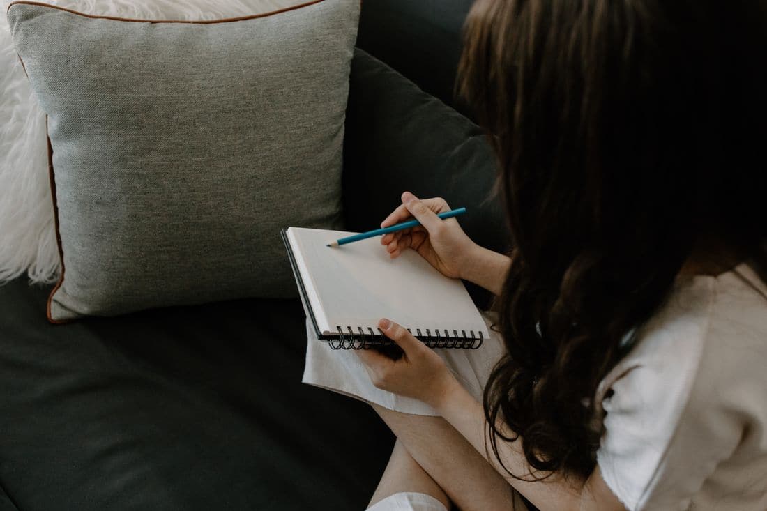 A woman sitting on a dark sofa writing in a spiral notepad with a blue pencil beside a grey cushion