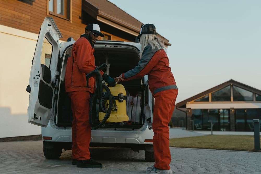 Two cleaners loading equipment into a van outside a building