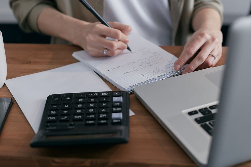 Hands writing figures beside a calculator and a laptop