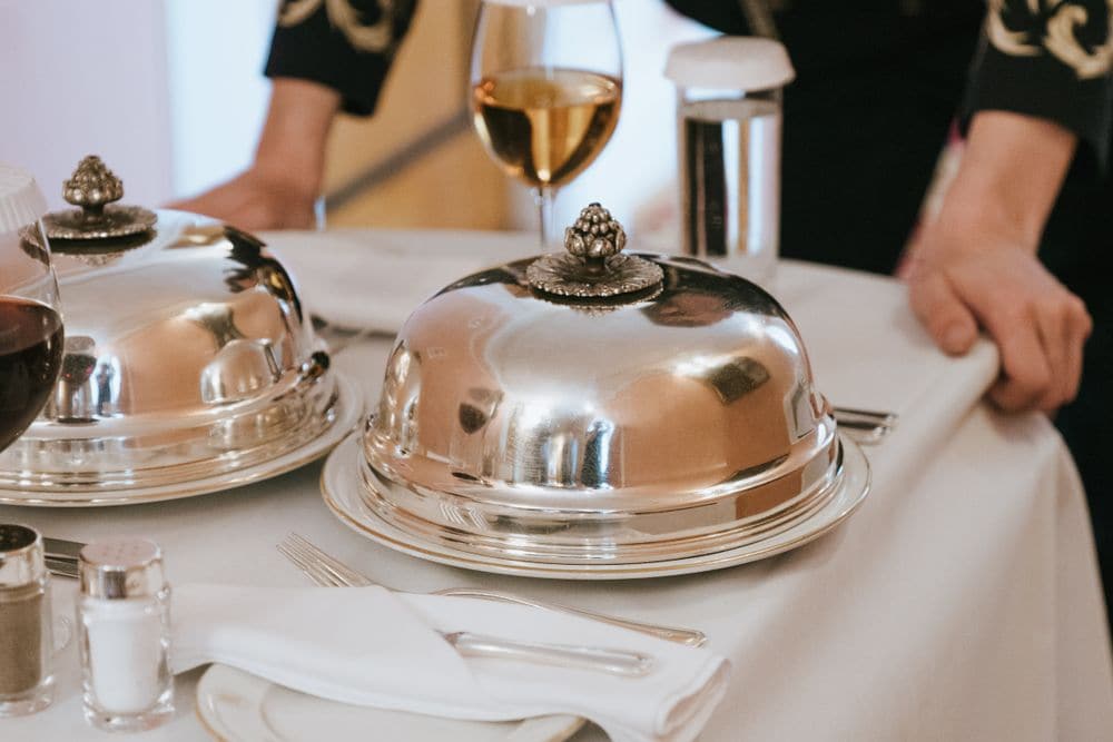 A waiter setting a table with covered silver dishes