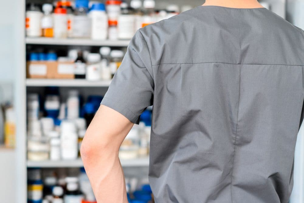 A pharmacy worker in scrubs facing shelves of medicines